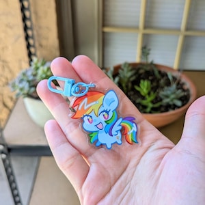 May include: A clear acrylic keychain featuring a cartoon pony character with a light blue body, a rainbow-colored mane and tail, and a lightning bolt symbol on its forehead. The keychain includes a light blue plastic lobster clasp attachment.