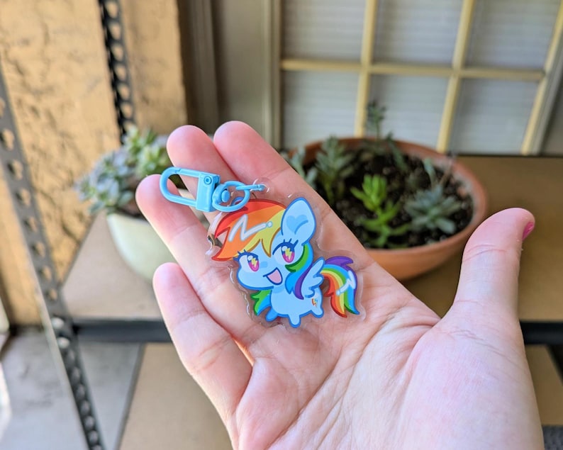 May include: A clear acrylic keychain featuring a cartoon pony character with a light blue body, a rainbow-colored mane and tail, and a lightning bolt symbol on its forehead. The keychain includes a light blue plastic lobster clasp attachment.