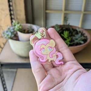May include: A clear acrylic keychain featuring a cartoon pony with a light yellow body, long flowing pink hair, and large blue eyes. The character has small wings and a light green metal lobster clasp attached to the top of the transparent charm.