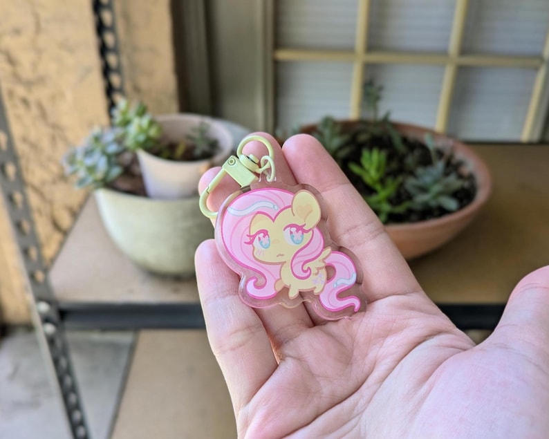 May include: A clear acrylic keychain featuring a cartoon pony with a light yellow body, long flowing pink hair, and large blue eyes. The character has small wings and a light green metal lobster clasp attached to the top of the transparent charm.