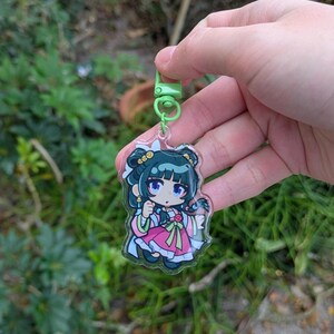 K&ouml;nnte beinhalten: Ein klarer Acryl-Schl&uuml;sselanh&auml;nger mit einer Cartoon-Figur in einem rosa und wei&szlig;en Outfit mit gr&uuml;nen Akzenten. Der Schl&uuml;sselanh&auml;nger hat einen gr&uuml;nen Verschluss und einen silbernen Schl&uuml;sselring. Die Figur hat dunkles Haar und blaue Augen. Der Hintergrund ist verschwommenes gr&uuml;nes Laub.