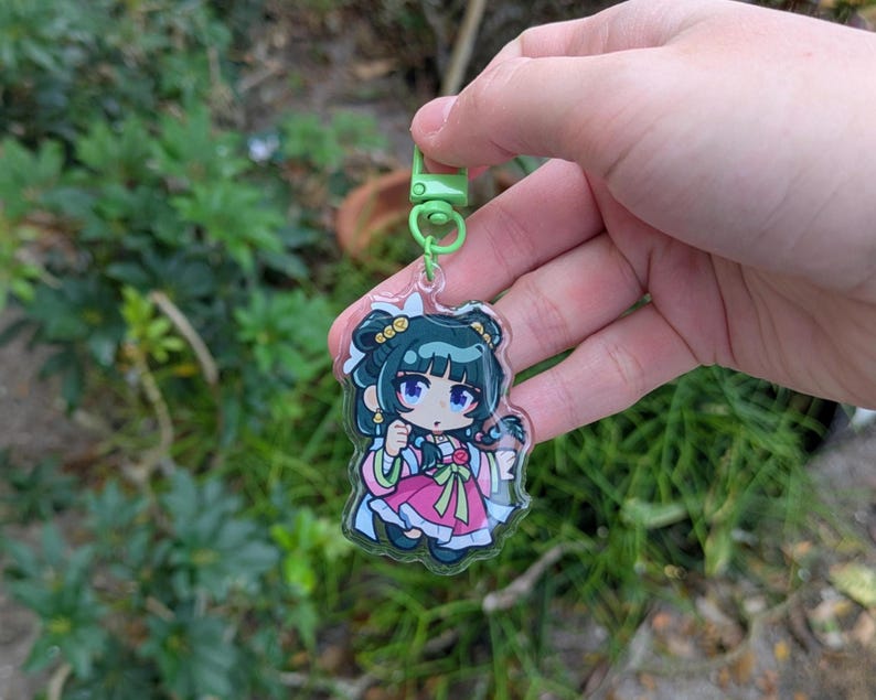 K&ouml;nnte beinhalten: Ein klarer Acryl-Schl&uuml;sselanh&auml;nger mit einer Cartoon-Figur in einem rosa und wei&szlig;en Outfit mit gr&uuml;nen Akzenten. Der Schl&uuml;sselanh&auml;nger hat einen gr&uuml;nen Verschluss und einen silbernen Schl&uuml;sselring. Die Figur hat dunkles Haar und blaue Augen. Der Hintergrund ist verschwommenes gr&uuml;nes Laub.