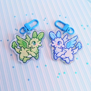 May include: Two acrylic dragon keychains. One is green and yellow, the other is blue and white. Each keychain has a blue clasp. The dragons have wings and are on a light blue background with a striped pattern and scattered star-shaped confetti.