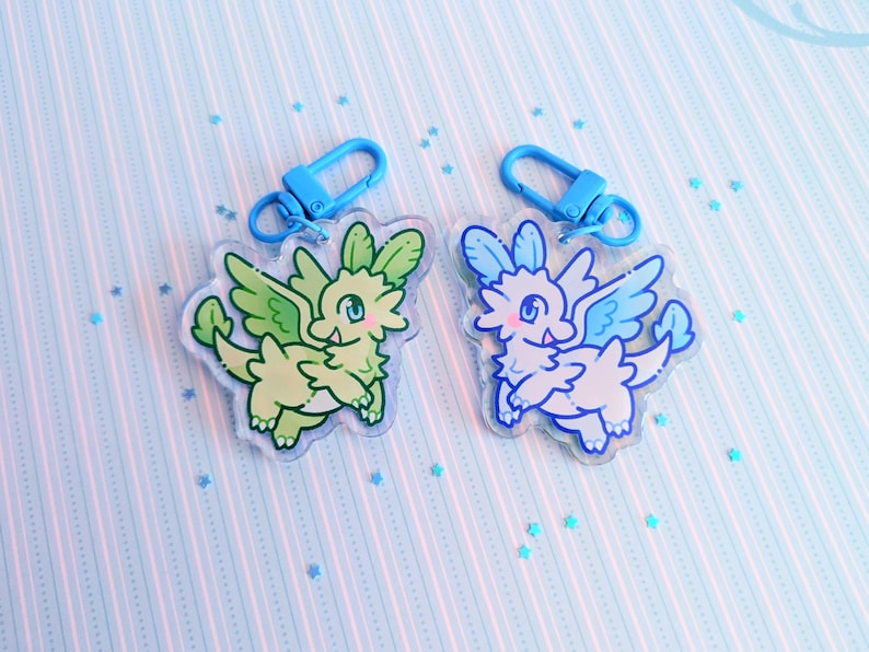 May include: Two acrylic dragon keychains. One is green and yellow, the other is blue and white. Each keychain has a blue clasp. The dragons have wings and are on a light blue background with a striped pattern and scattered star-shaped confetti.