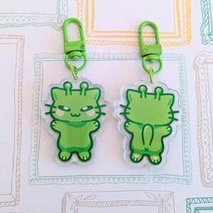 May include: Two green alien cat keychains with green eyes and a green body. The keychains are attached to green metal key rings.