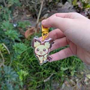 May include: A clear acrylic keychain featuring a cartoon Pichu character with yellow and brown accents. The keychain has a yellow clasp and ring. The background is blurred with green foliage.