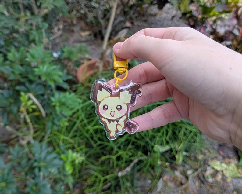 May include: A clear acrylic keychain featuring a cartoon Pichu character with yellow and brown accents. The keychain has a yellow clasp and ring. The background is blurred with green foliage.