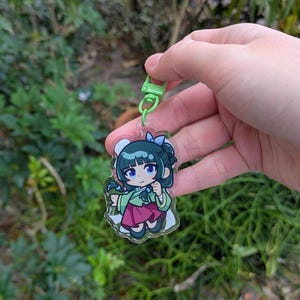K&ouml;nnte beinhalten: Ein klarer Acryl-Anh&auml;nger mit einem Cartoon-M&auml;dchen in einem traditionellen koreanischen Hanbok. Der Anh&auml;nger hat einen gr&uuml;nen Verschluss und eine blaue Schleife. Das M&auml;dchen hat schwarzes Haar und tr&auml;gt ein gr&uuml;nes Oberteil und einen roten Rock. Der Hintergrund ist unscharf.