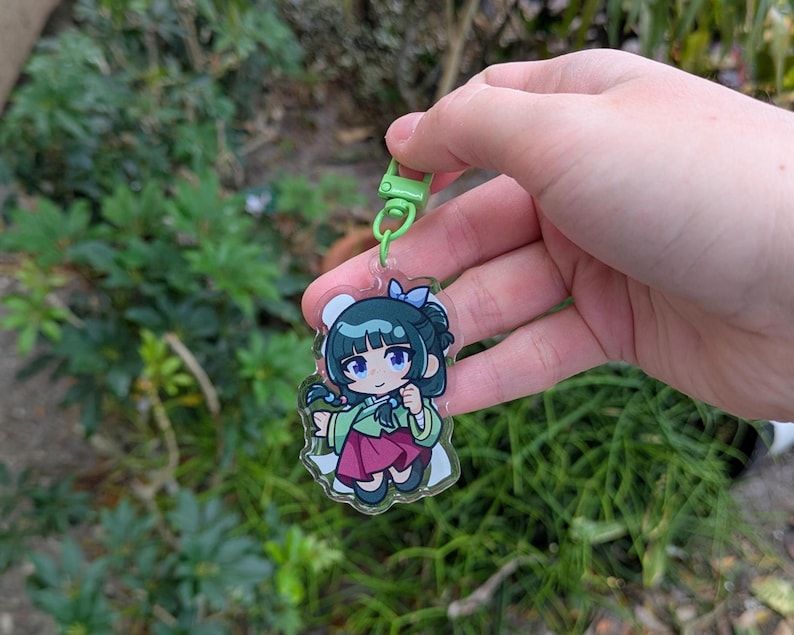 K&ouml;nnte beinhalten: Ein klarer Acryl-Anh&auml;nger mit einem Cartoon-M&auml;dchen in einem traditionellen koreanischen Hanbok. Der Anh&auml;nger hat einen gr&uuml;nen Verschluss und eine blaue Schleife. Das M&auml;dchen hat schwarzes Haar und tr&auml;gt ein gr&uuml;nes Oberteil und einen roten Rock. Der Hintergrund ist unscharf.