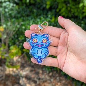 May include: A blue cat-shaped keychain with a gold-colored clasp. The cat has yellow eyes, a pink mouth, and blue stripes. The keychain is held in a hand against a blurred green background.