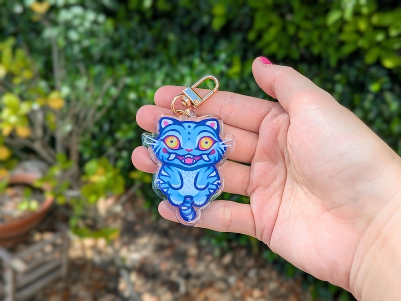 May include: A blue cat-shaped keychain with a gold-colored clasp. The cat has yellow eyes, a pink mouth, and blue stripes. The keychain is held in a hand against a blurred green background.