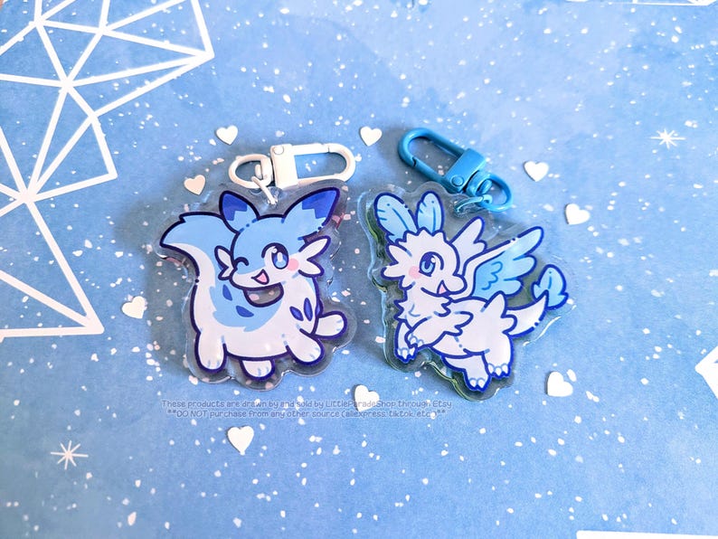 May include: Two acrylic keychains featuring cartoon-style creatures in shades of blue and white. One keychain has a white clasp, and the other has a blue clasp. The background is light blue with white geometric shapes and heart-shaped confetti.