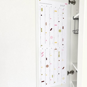 K&ouml;nnte beinhalten: Ein vertikaler Wandkalender f&uuml;r das Jahr 2027 auf wei&szlig;em Papier mit zw&ouml;lf Spalten f&uuml;r jeden Monat. Das Design zeigt verschiedene geometrische Formen in Rosa, Gelb, Braun und Olivgr&uuml;n, die &uuml;ber das Raster verteilt sind, mit dem Text 2027 in der oberen rechten Ecke.