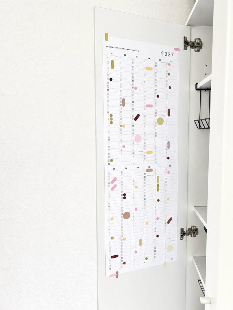 K&ouml;nnte beinhalten: Ein vertikaler Wandkalender f&uuml;r das Jahr 2027 auf wei&szlig;em Papier mit zw&ouml;lf Spalten f&uuml;r jeden Monat. Das Design zeigt verschiedene geometrische Formen in Rosa, Gelb, Braun und Olivgr&uuml;n, die &uuml;ber das Raster verteilt sind, mit dem Text 2027 in der oberen rechten Ecke.