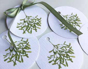 Könnte beinhalten: Fünf weiße runde Geschenkanhänger mit einer grünen Mistelzweig-Illustration. Jeder Anhänger hat ein Loch oben und ein grünes Satinband ist um einen der Anhänger gebunden.