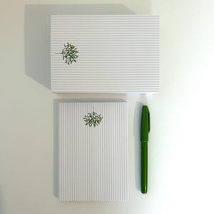 May include: Two white notepads with gray horizontal stripes and a green sprig design, one stacked on top of the other. A green pen is positioned vertically next to the bottom notepad. The items are on a white surface.