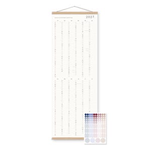 K&ouml;nnte beinhalten: Ein vertikaler Wandkalender f&uuml;r 2027 auf wei&szlig;em Papier mit schwarzem Text, gehalten von hellen Holzleisten oben und unten mit einer Schnur zum Aufh&auml;ngen. Ein separates rechteckiges Blatt mit kleinen, runden Aufklebern in Farbverl&auml;ufen liegt bei.