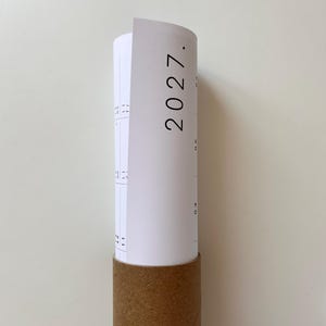 K&ouml;nnte beinhalten: Ein wei&szlig;er Papierkalender f&uuml;r das Jahr 2027, der teilweise aufgerollt und in eine zylindrische Versandrolle aus brauner Pappe gesteckt ist. Der Kalender zeigt schwarze Drucktexte und Gitterlinien, wobei das Jahr 2027 deutlich sichtbar ist.