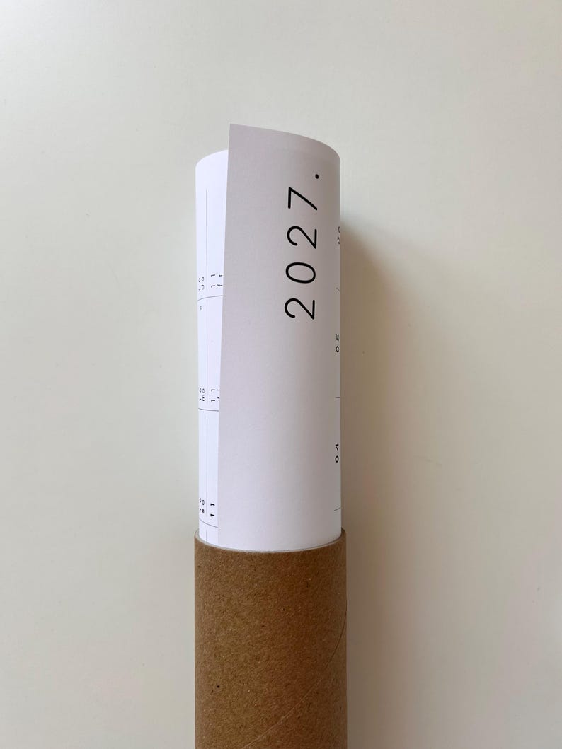 K&ouml;nnte beinhalten: Ein wei&szlig;er Papierkalender f&uuml;r das Jahr 2027, der teilweise aufgerollt und in eine zylindrische Versandrolle aus brauner Pappe gesteckt ist. Der Kalender zeigt schwarze Drucktexte und Gitterlinien, wobei das Jahr 2027 deutlich sichtbar ist.