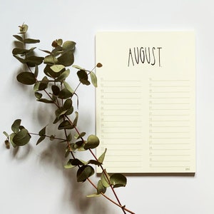 May include: A cream-coloured notepad with a handwritten calendar for the month of August. The notepad has a green line running down the centre and green lines separating the days of the month. The notepad is on a white surface with a sprig of eucalyptus leaves to the left.