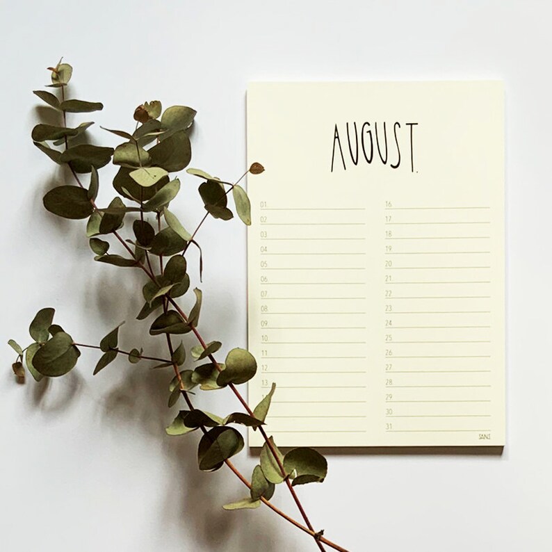 May include: A cream-coloured notepad with a handwritten calendar for the month of August. The notepad has a green line running down the centre and green lines separating the days of the month. The notepad is on a white surface with a sprig of eucalyptus leaves to the left.