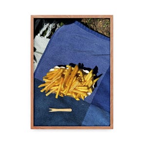 Könnte beinhalten: Gerahmtes Bild einer Picknick-Szene. Ein Papierbehälter mit goldbraunen Pommes frites liegt auf einer blauen Decke. Eine kleine Holzgabel liegt daneben. Die Szene ist in einem Holzrahmen gerahmt.