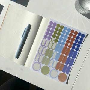 K&ouml;nnte beinhalten: Ein offenes Notizbuch mit linierten Seiten und einem hellblauen Druckkugelschreiber auf der linken Seite. Rechts daneben befindet sich ein Aufkleberbogen mit verschiedenen Formen, darunter kleine Kreise und abgerundete Rechtecke in Lila, Oliv, Blau und Orange.