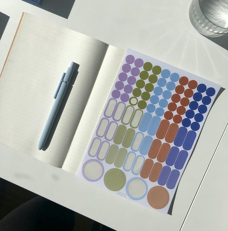 K&ouml;nnte beinhalten: Ein offenes Notizbuch mit linierten Seiten und einem hellblauen Druckkugelschreiber auf der linken Seite. Rechts daneben befindet sich ein Aufkleberbogen mit verschiedenen Formen, darunter kleine Kreise und abgerundete Rechtecke in Lila, Oliv, Blau und Orange.