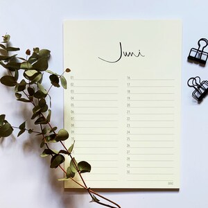 May include: A blank monthly calendar page with the word "Juni" written at the top. The calendar is on a white surface with a sprig of eucalyptus leaves and two black binder clips.