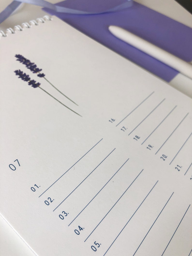 Pu&ograve; includere: Un taccuino bianco a spirale con linee blu e numeri. Il taccuino ha un fiore di lavanda pressato sulla copertina.