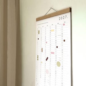 K&ouml;nnte beinhalten: Ein Wandkalender f&uuml;r das Jahr 2027 auf wei&szlig;em Papier mit vertikalem Raster. Der Kalender zeigt kleine, bunte geometrische Formen, die &uuml;ber die Daten verteilt sind, und wird oben von einer hellbraunen magnetischen Holzleiste mit schwarzer Schnur gehalten.