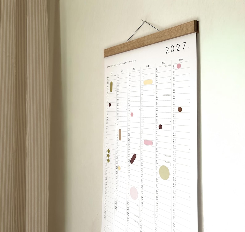 K&ouml;nnte beinhalten: Ein Wandkalender f&uuml;r das Jahr 2027 auf wei&szlig;em Papier mit vertikalem Raster. Der Kalender zeigt kleine, bunte geometrische Formen, die &uuml;ber die Daten verteilt sind, und wird oben von einer hellbraunen magnetischen Holzleiste mit schwarzer Schnur gehalten.