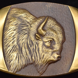 May include: A gold-toned belt buckle with a detailed, embossed image of a bison's head. The bison's fur is textured and the buckle has a smooth, polished finish.