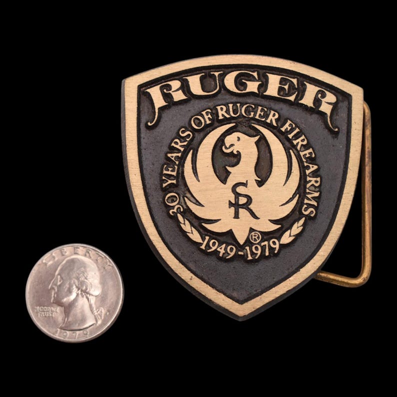 May include: A vintage metal belt buckle with the word "RUGER" at the top. The buckle features a shield design with the text "30 YEARS OF RUGER FIREARMS 1949-1979" and a bird emblem. A quarter is next to the buckle.