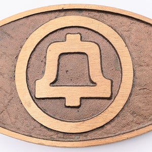 May include: A brown metal belt buckle with a raised, circular design featuring a bell silhouette.