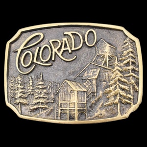 May include: A rectangular, bronze-toned belt buckle with the word "Colorado" in a cursive font. The buckle features a detailed relief of a mountain scene with buildings and trees.