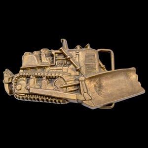 May include: A detailed, gold-colored belt buckle shaped like a bulldozer. The buckle features intricate detailing of the vehicle's tracks, blade, and engine components. The design is set against a black background.