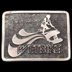 May include: Rectangular silver-colored belt buckle with a textured background. The design features a person riding a jet ski, with the word "WETBIKE" in bold letters below. The buckle has a dark background.
