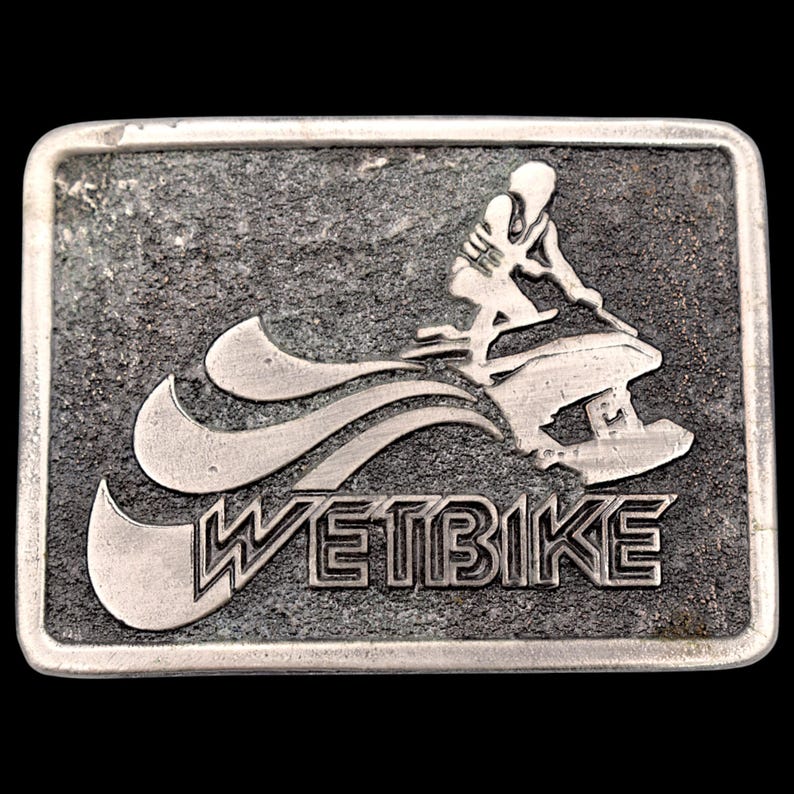 May include: Rectangular silver-colored belt buckle with a textured background. The design features a person riding a jet ski, with the word "WETBIKE" in bold letters below. The buckle has a dark background.