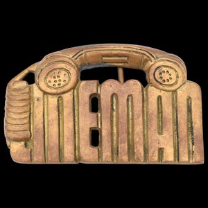 May include: A vintage brass belt buckle with the word "MEMAN" in bold letters. The buckle features a telephone handset design above the letters, with a textured surface and a classic, retro aesthetic.