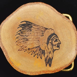 May include: A wooden belt buckle with a Native American chief design. The buckle is made from a slice of wood, featuring a detailed black and brown engraved profile of a chief wearing a feathered headdress. The buckle has a gold-colored metal clasp.