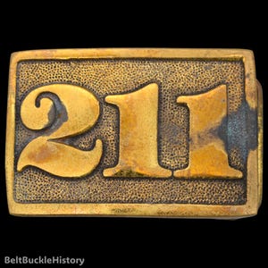 May include: A rectangular brass belt buckle featuring the raised number 211 in a bold, serif font. The surface of the buckle has a textured, stippled background with a weathered, golden-brown patina and visible signs of oxidation along the edges.