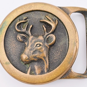May include: A brass belt buckle with a detailed, embossed image of a deer head with antlers. The buckle is circular with a raised rim and a smooth, polished finish.