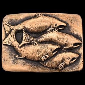 May include: A rectangular bronze-colored belt buckle featuring a relief design of four fish swimming in the same direction. The fish are detailed with fins, scales, and open mouths, set against a textured background.