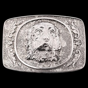 May include: A rectangular silver-colored belt buckle with a detailed relief of a dog's face in the center. The dog is surrounded by a circular frame and decorative floral elements. The buckle has a textured background.