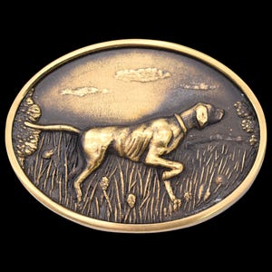 May include: Oval-shaped belt buckle with a detailed relief of a dog in a field. The dog is golden-colored, set against a dark background with a golden border. The dog is running through tall grass.