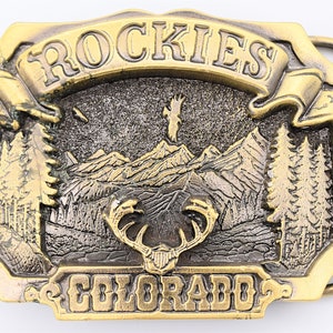 May include: A gold-tone belt buckle with a raised design of a mountain range, a deer head, and the words "ROCKIES COLORADO".