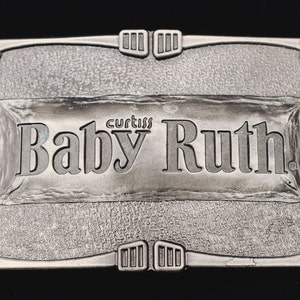May include: A silver-colored metal belt buckle with the words "Baby Ruth" in a rectangular frame. The brand name "Curtiss" is above the text. The buckle has an ornate border with decorative elements.