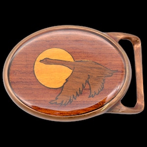 May include: Oval belt buckle with a wooden inlay design. The buckle features a flying goose silhouette against a yellow sun, set on a background of dark wood. The buckle has a bronze-colored metal frame and clasp.