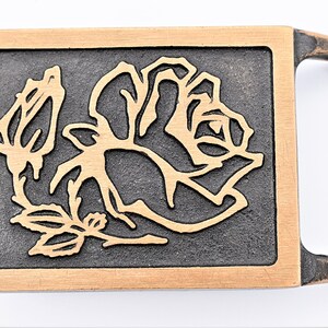 May include: A rectangular belt buckle with a rose design. The buckle features a dark gray background with a gold-colored rose and a gold-colored border. The handle is a darker, aged gold color.